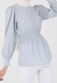 Vaaleansininen pusero, jossa on hunajakennokuvioitu vyötärö, puffimaiset pitkät hihat ja teksturoitu kangas. Tyylitelty valkoisten housujen kanssa, joissa on pyöristetty pääntie.