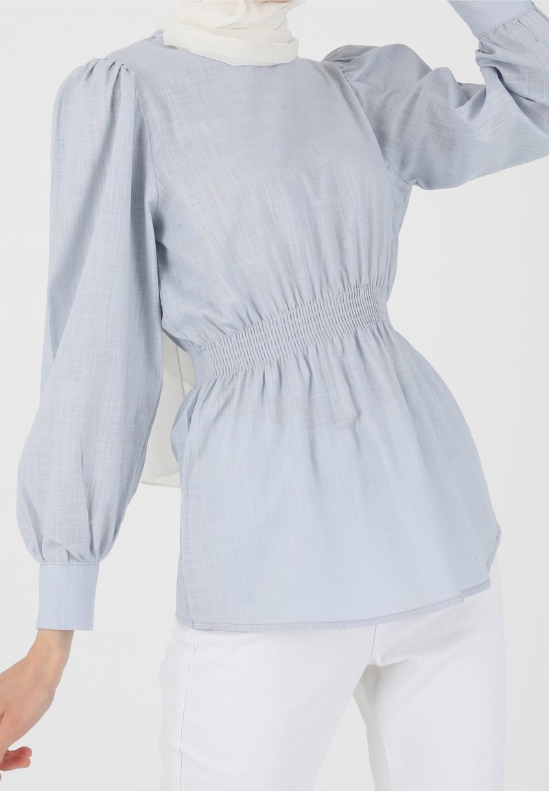 Vaaleansininen pusero, jossa on hunajakennokuvioitu vyötärö, puffimaiset pitkät hihat ja teksturoitu kangas. Tyylitelty valkoisten housujen kanssa, joissa on pyöristetty pääntie.