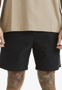Shorts en velours côtelé noir avec une finition texturée, une taille standard et des poches latérales, associés à un t-shirt beige clair.