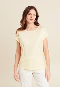 Lichtgele t-shirt met korte mouwen en een subtiele glans, ronde halslijn en opgerolde mouwmanchetten, gecombineerd met crèmekleurige broek.
