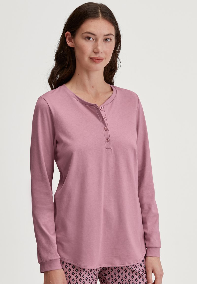 Langarmige roze top met een knoopsluiting bij de hals. Zachte textuur, relaxte pasvorm en contrasterende manchetten. Gecombineerd met een pantalon met een patroon.