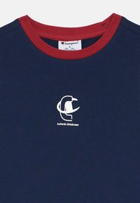 Μπλε ναυτικό T-shirt με κόκκινο ριμπ ζωνάρι στο γιακά, με λευκό λογότυπο και κείμενο "Authentic Athleticwear" στο μπροστινό μέρος. Απαλό βαμβακερό υλικό.