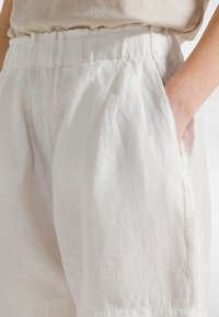 Białe lniane spodnie o luźnym kroju, z elastycznym pasem w talii i bocznymi kieszeniami. Tkanina ma subtelną teksturę i lekki połysk.