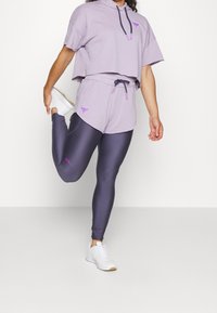 Žena se protahuje, má na sobě levandulovou mikinu, odpovídající šortky, tmavě fialové legíny a bílé tenisky na bílém pozadí.