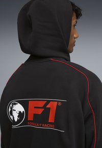 Sort hættebluse med rød accent og hvid piping, som har et stort F1-logo og verdenskortgrafik på ryggen. Blødt stof.