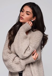 Beige gestrickter Pullover mit lockeren, übergroßen Ärmeln und einem strukturierten Muster. Das Model hält eine Hand an ihr Kinn und präsentiert Ohrstecker und einen Ring.