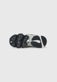 Semelle en caoutchouc noir de baskets présentant des motifs de crampons variés, des rainures centrales et une semelle intermédiaire blanche. Le design comprend des marquages de la marque et des détails texturés.
