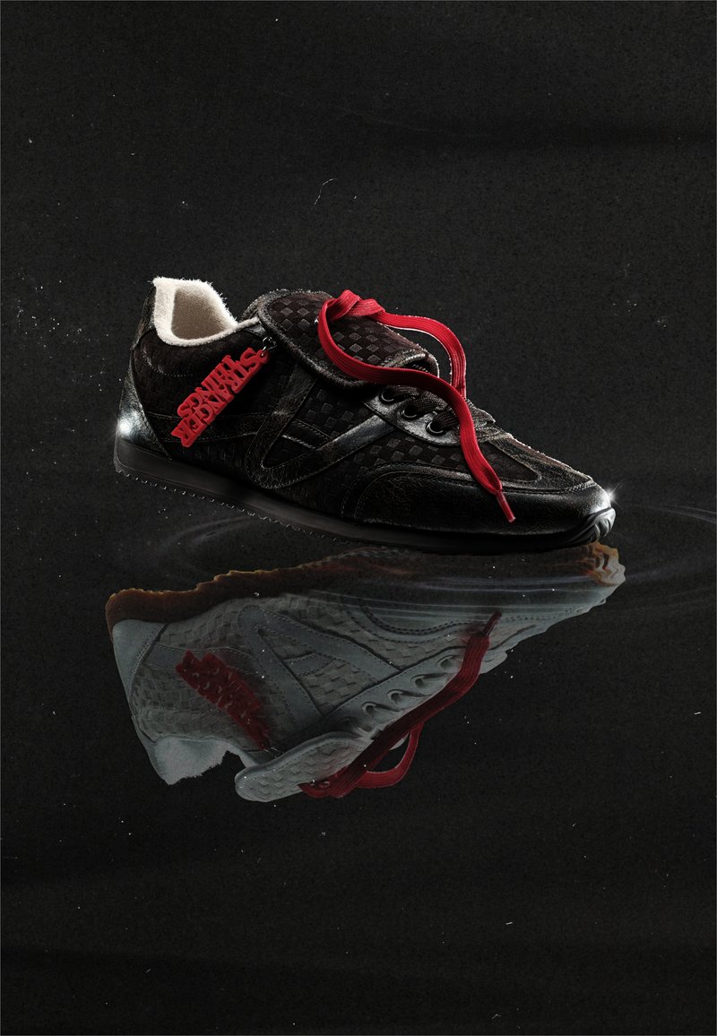 Black sneaker with red laces and a red "STRANGER THINGS" tag, reflecting on a glossy dark surface with a subtle ripple effect.