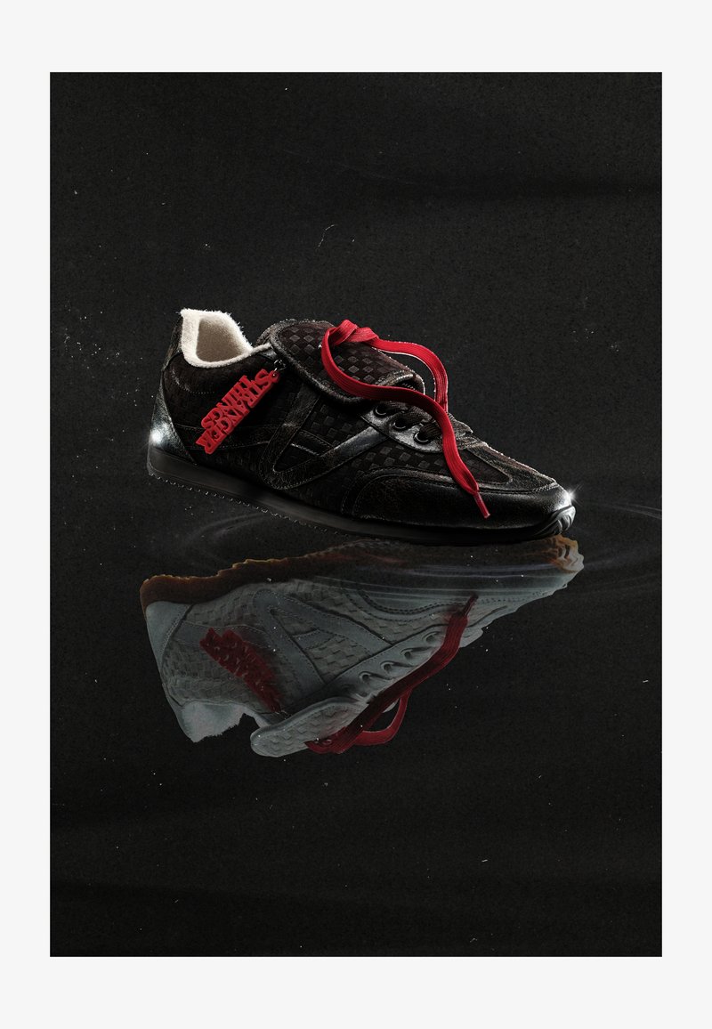 Black sneaker with red laces and a red "STRANGER THINGS" tag, reflecting on a glossy dark surface with a subtle ripple effect.