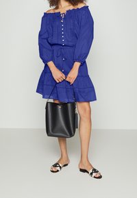 Off-shoulder blå klänning med knappdetaljer och dragsko i midjan; matchas med en svart bucket bag och mönstrade sandaler.