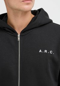Zwarte zip-up hoodie met een zilveren rits en een witte "A.R.C." opdruk op de bovenkant van de borst. Zacht materiaal met een textuur en een capuchon.