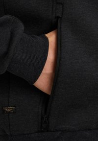 Sweatshirt noir avec une poche zippée et des poignets côtelés. Texture douce avec un motif subtil. L'étiquette indique "reborn unlimited."