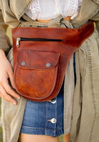 Brown leather waist bag with a front zipper pocket and a flap pocket, featuring black hardware and a textured finish, worn over a denim skirt.