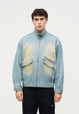 Młody mężczyzna z krótkimi kręconymi włosami, ubrany w jasnoniebieską jeansową kurtkę na zamek z poplamionymi przednimi kieszeniami i czarne spodnie.