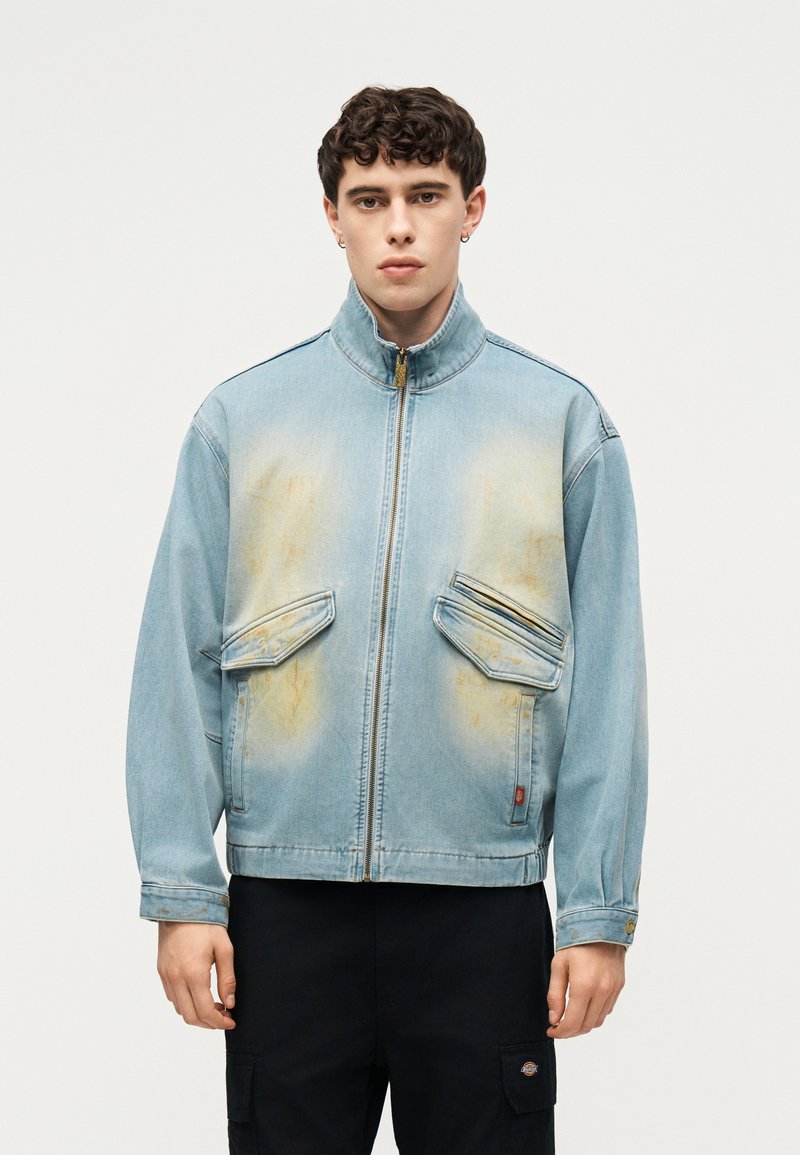 Νεαρός με κοντά σγουρά μαλλιά που φοράει ανοιχτό μπλε τζιν μπουφάν με φερμουάρ και λεκιασμένες μπροστινές τσέπες και μαύρο παντελόνι.