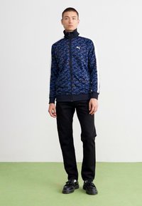 Blå zip-up jakke med mønsterdesign, høj krave, hvide sidelister, kombineret med sorte bukser og sko. Glat tekstur, afslappet stil.