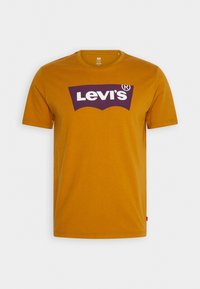 T-shirt z krótkim rękawem w kolorze musztardowym, wykonany z bawełny, z dużym fioletowym logo Levi's w klasycznym łukowatym wzorze na przodzie.