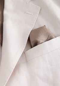 Blazer en lin beige avec une finition texturée et un mouchoir de poche en taupe clair, arborant un motif subtil.