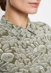 Camicia verde con un motivo floreale bianco, colletto e dettagli con bottoni. Il tessuto sembra morbido e testurizzato.