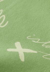 Svetlozelená látka s bielym handwritten textom a krížovými vzormi, s jemnou textúrou a subtílnym tkaným vzhľadom.