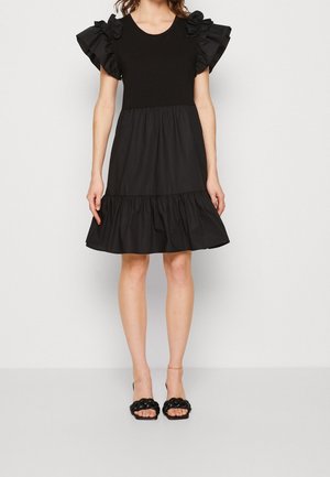 Vrouw draagt een zwarte jurk tot op de knie met gerimpelde mouwtjes en een roklagen, gecombineerd met zwarte hakken met gevlochten bandjes.