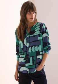 Flerfärgad blus med en blandning av geometriska former i grönt, marinblått och teal, med V-ringning och långa ärmar. Tillverkad av ett lättviktsmaterial.