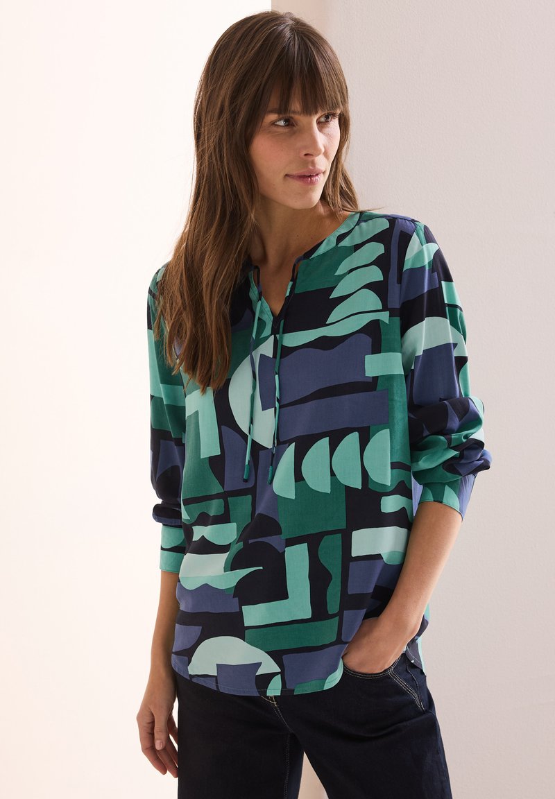 Flerfärgad blus med en blandning av geometriska former i grönt, marinblått och teal, med V-ringning och långa ärmar. Tillverkad av ett lättviktsmaterial.