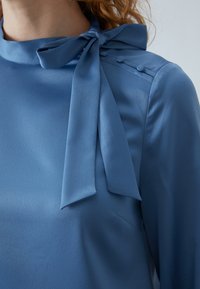 Világoskék blúz, amelyet masnis nyak jellemez, sima textúrával és díszítő gombokkal a vállon. Elegáns dizájn, puha redőzéssel.