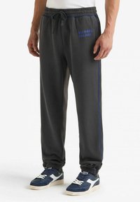 Pantaloni della tuta grigi realizzati in materiale morbido con strisce laterali blu e ricami. Presentano un elastico in vita con cordoncino e polsini alle caviglie.