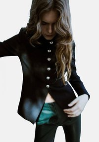 Zwarte getailleerde blazer met zilveren knopen en een hoge kraag, gecombineerd met donkergroene broeken met een getailleerde snit en subtiele glans.