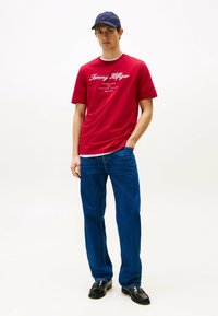 Rød kortærmet t-shirt med hvidt "Tommy Hilfiger" logo, sat sammen med blå lige ben jeans og sorte lædersko, toppet med en marineblå cap.