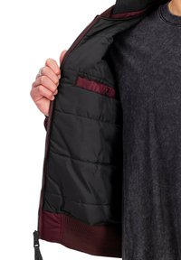 Bordeauxfarbene Jacke mit schwarzem Innenfutter, mit einem gesteppte Innenfutter, Reißverschluss und seitlichen Taschen, offen präsentiert.