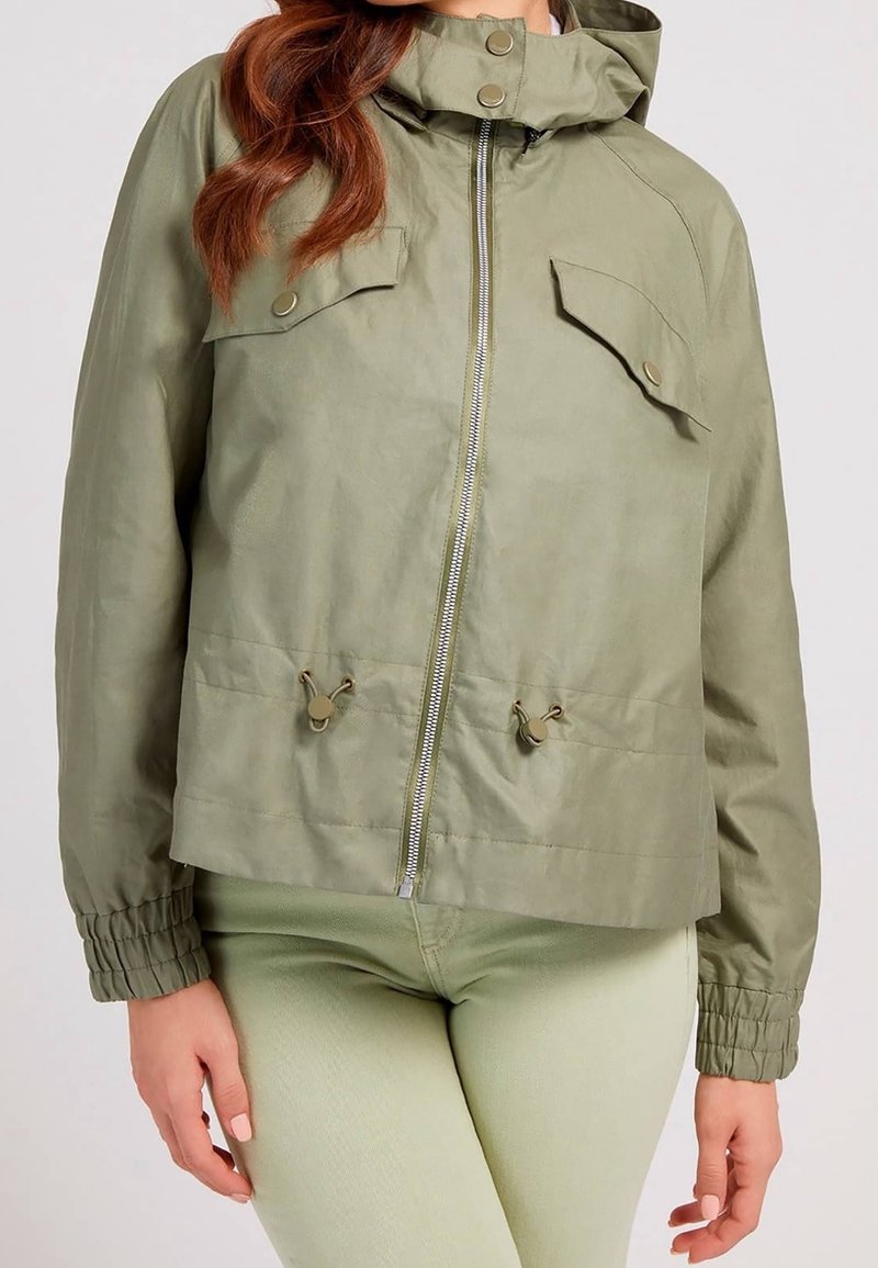 Veste à capuche vert olive clair avec fermeture éclair à l'avant, poches à rabat et cordons réglables, portée avec un pantalon vert pâle.