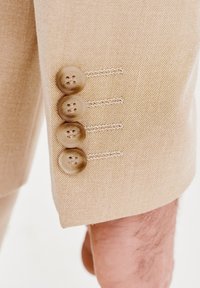 Rękaw beżowej marynarki z czterema zaokrąglonymi, jasnobrązowymi guzikami. Teksturowany materiał z delikatnym wzorem, dekoracyjne przeszycia wzdłuż guzików.