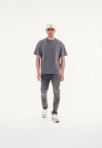 Camiseta de algodón gris con mangas cortas, combinada con unos jeans grises desgastados. El modelo lleva una gorra blanca y zapatillas blancas. Diseño simple, con un mínimo de detalles.