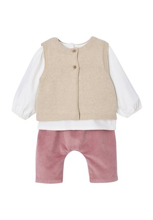 Beige gestrickte Weste mit zwei Knöpfen, die über einem weißen Langarmshirt getragen wird, kombiniert mit pinken Cordhose mit abgerundetem Saum.