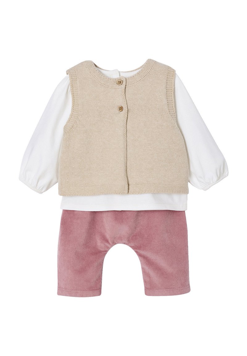Beige gestrickte Weste mit zwei Knöpfen, die über einem weißen Langarmshirt getragen wird, kombiniert mit pinken Cordhose mit abgerundetem Saum.