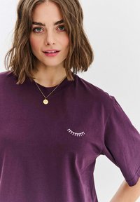 Fialové bavlněné tričko s jednoduchým designem řas v bílé barvě na levé straně. Krátké rukávy, kulatý výstřih, hladká textura.