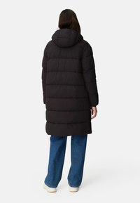 Person standing, wearing a long black hooded puffer coat, blue jeans, and white shoes, shown from the back against a plain background.