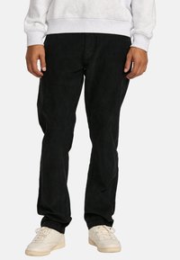 Black corduroy pants with a relaxed fit. Features a textured surface, side pockets, and worn with white sneakers and a gray sweatshirt.