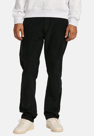 Black corduroy pants with a relaxed fit. Features a textured surface, side pockets, and worn with white sneakers and a gray sweatshirt.
