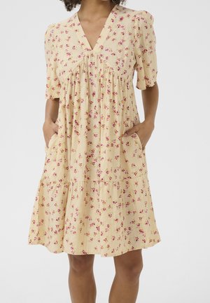 Femme portant une robe jaune clair longueur genou avec un imprimé floral rose, des manches courtes bouffantes, un col en V et les mains dans les poches latérales.