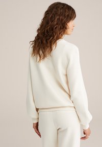 Mujer con cabello castaño ondulado, vistiendo una blusa de manga larga color crema y pantalones a juego, de perfil contra un fondo neutro.