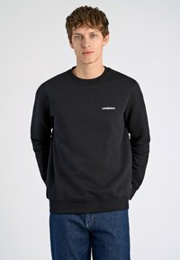 Svart sweatshirt i mjukt tyg, med rund halsringning, långa ärmar och en liten vit "LINDBERGH"-logotyp på vänster bröst. Bärs med blå jeans.