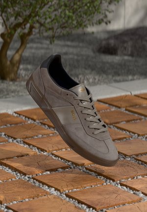 Sneaker in camoscio marrone con colletto nero e logo "BOSS" dorato, che fluttua sopra una mattonella in legno e una superficie esterna di ghiaia.
