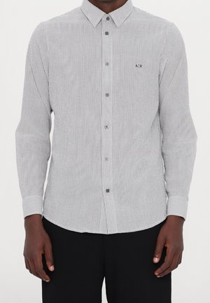 Mężczyzna noszący białą, dopasowaną koszulę z długim rękawem, zapinaną na guziki, z cienkimi czarnymi pionowymi paskami oraz czarne spodnie.