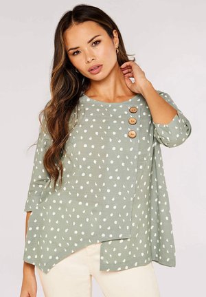 Frau mit langem, welligem Haar, die eine hellgrüne Bluse mit weißen Punkten und drei hölzernen Knöpfen an der Schulter trägt und mit der Hand nahe am Hals posiert.