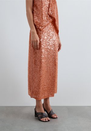 Mujer con falda midi de lentejuelas color melocotón brillante y top a juego, combinados con sandalias negras de tacón sobre un fondo de suelo gris.