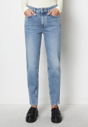 Persona que lleva puestos unos vaqueros rectos de color azul claro y mocasines negros con suela gruesa, de pie con las manos en los bolsillos frente a una pared blanca.