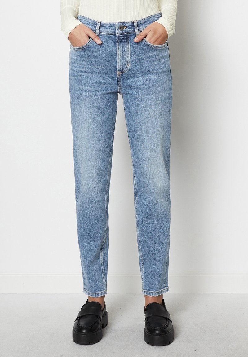 Personne portant un jean droit bleu clair et des mocassins noirs épais, debout les mains dans les poches contre un mur blanc.